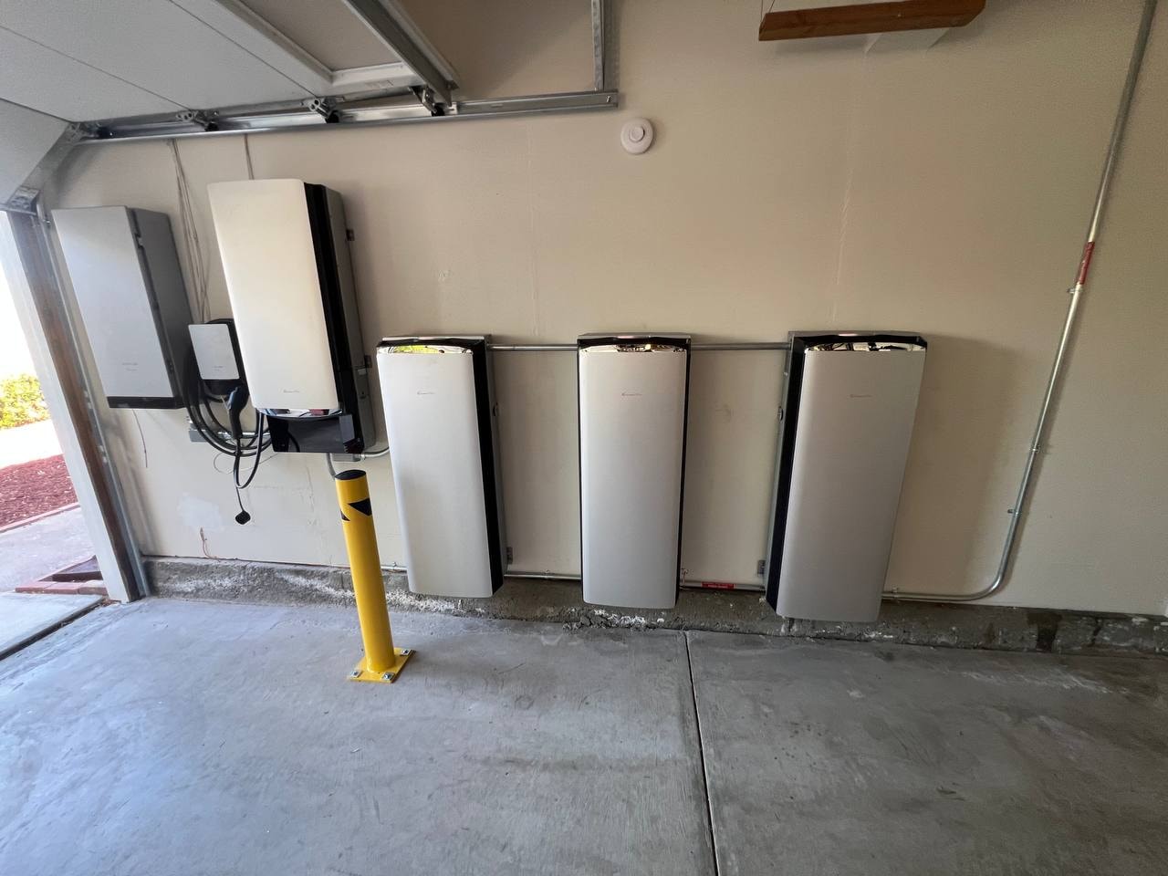 Solar inverter and three battery units mounted on a garage wall, with a yellow bollard in front protecting the equipment from vehicle contact