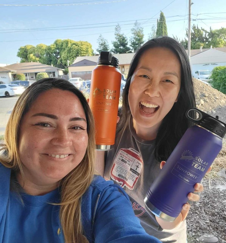 Two smiling women take a selfie outdoors in a residential neighborhood with houses, cars, trees, and power lines in the background. The woman on the left has long brown hair, wears a blue shirt and hoodie, and is smiling warmly. The woman on the right has long black hair, wears a gray t-shirt with a red and white patch, and is laughing with her mouth open. They are both holding insulated water bottles branded with 'Solar Survey Team' and 'SunPower'—one orange and one purple.