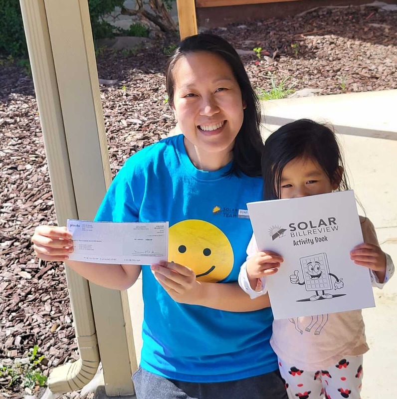 A person wearing a blue t-shirt with a large yellow smiley face and the text 'SOLAR BILL TEAM' is holding a check. Next to the person, a child is holding a booklet titled 'SOLAR BILL REVIEW Activity Book' with an illustration of a smiling solar panel giving a thumbs up. The background shows an outdoor setting with mulch and a concrete path.