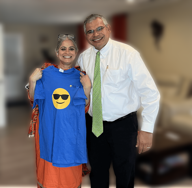 A smiling woman in a red and orange traditional outfit holds up a blue T-shirt featuring a sunglasses emoji. She is standing next to a man wearing a white dress shirt, a green tie, and black pants, who is also smiling. They are posing together in a warmly lit living room with beige walls, a cream-colored sofa, a wooden coffee table with books and small objects, and red curtains in the background. The woman has gray hair tied back, and the man has short graying hair and glasses.