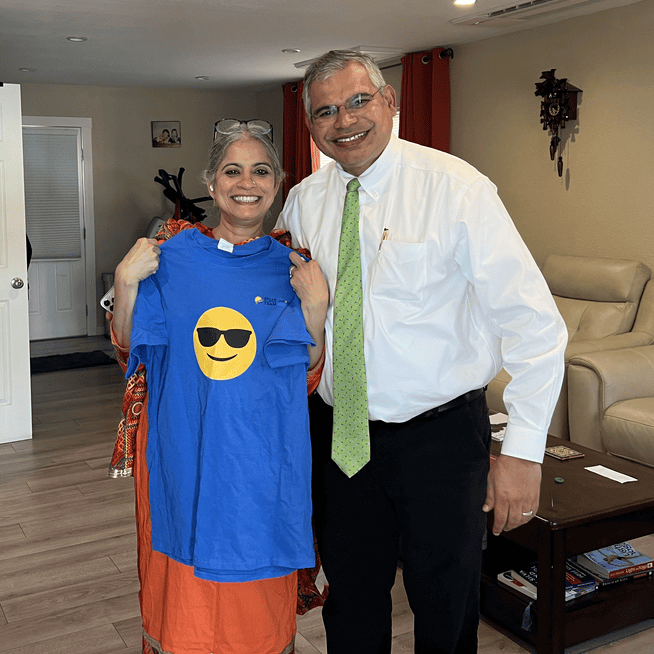 A smiling woman in a red and orange traditional outfit holds up a blue T-shirt featuring a sunglasses emoji. She is standing next to a man wearing a white dress shirt, a green tie, and black pants, who is also smiling. They are posing together in a warmly lit living room with beige walls, a cream-colored sofa, a wooden coffee table with books and small objects, and red curtains in the background. The woman has gray hair tied back, and the man has short graying hair and glasses.